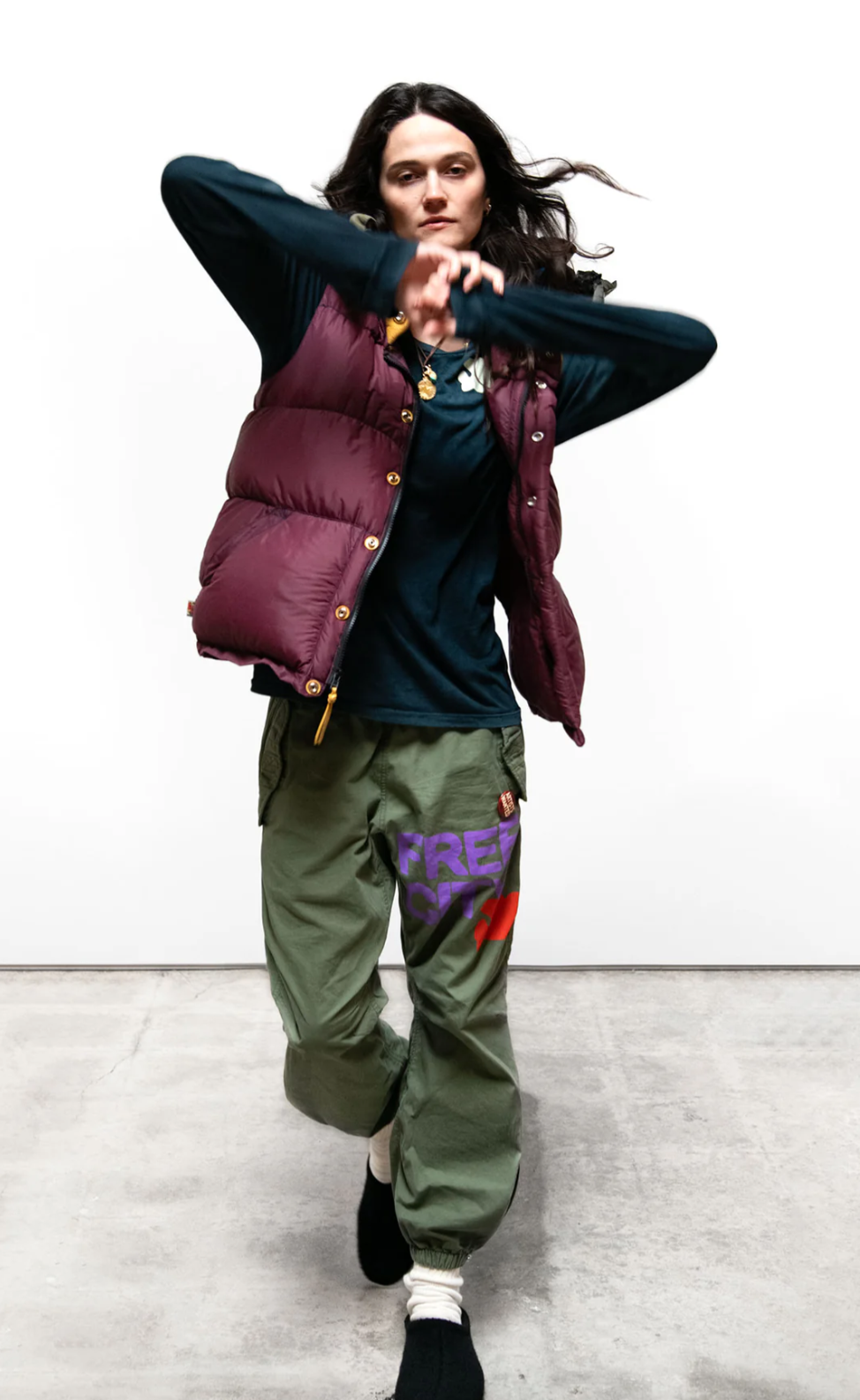 A person with long dark hair is dancing while wearing a burgundy puffy vest over a dark long-sleeved shirt and green RTU SPRVINT flap/snap LG BAG/SHELL pants from Free City (sparrow, LLC) that feature colorful text on one leg. They are standing against a plain white background on a concrete floor.