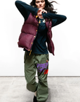 A person with long dark hair is dancing while wearing a burgundy puffy vest over a dark long-sleeved shirt and green RTU SPRVINT flap/snap LG BAG/SHELL pants from Free City (sparrow, LLC) that feature colorful text on one leg. They are standing against a plain white background on a concrete floor.