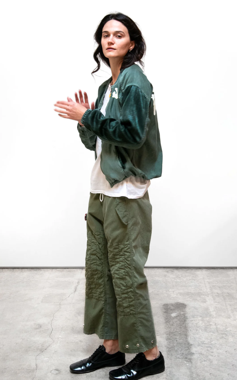 A person with long dark hair stands against a plain white background. They are wearing a green satin bomber jacket, a white shirt, RTU flap/snap KARATE BAG pant by Free City (sparrow, LLC) made from vintage army tents, and black shoes. Their hands are clasped together as they look forward.