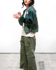 A person with long dark hair stands against a plain white background. They are wearing a green satin bomber jacket, a white shirt, RTU flap/snap KARATE BAG pant by Free City (sparrow, LLC) made from vintage army tents, and black shoes. Their hands are clasped together as they look forward.