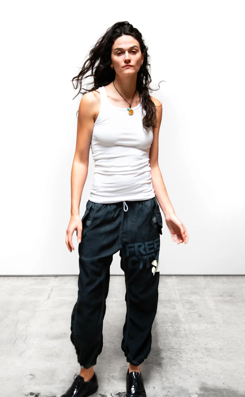 A woman with long, wavy hair stands against a white background, wearing a white tank top and the dark Flap/Snap LG MATTE/SATIN AirJump Pant from Free City (sparrow, LLC) with an elastic waist and the words "FREE CITY" on the left leg. Her black shoes complement her relaxed posture as her hair gently blows in the wind.