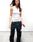 A woman with long, wavy hair stands against a white background, wearing a white tank top and the dark Flap/Snap LG MATTE/SATIN AirJump Pant from Free City (sparrow, LLC) with an elastic waist and the words "FREE CITY" on the left leg. Her black shoes complement her relaxed posture as her hair gently blows in the wind.