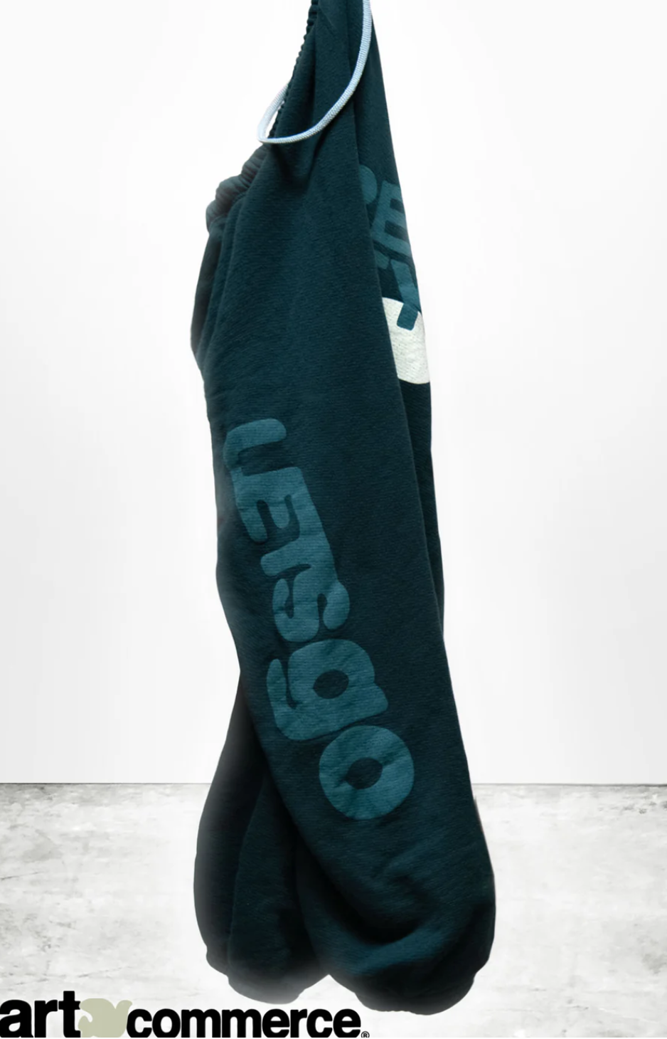 A dark green SUPERYUMM OG LETSGO sweats hoodie by Free City (sparrow, LLC) with "LET'S GO" written in large, light blue letters on the side. The oversized fit hoodie hangs against a plain white background, with the words "art commerce" and the FREECITY logo visible at the bottom left corner of the image.