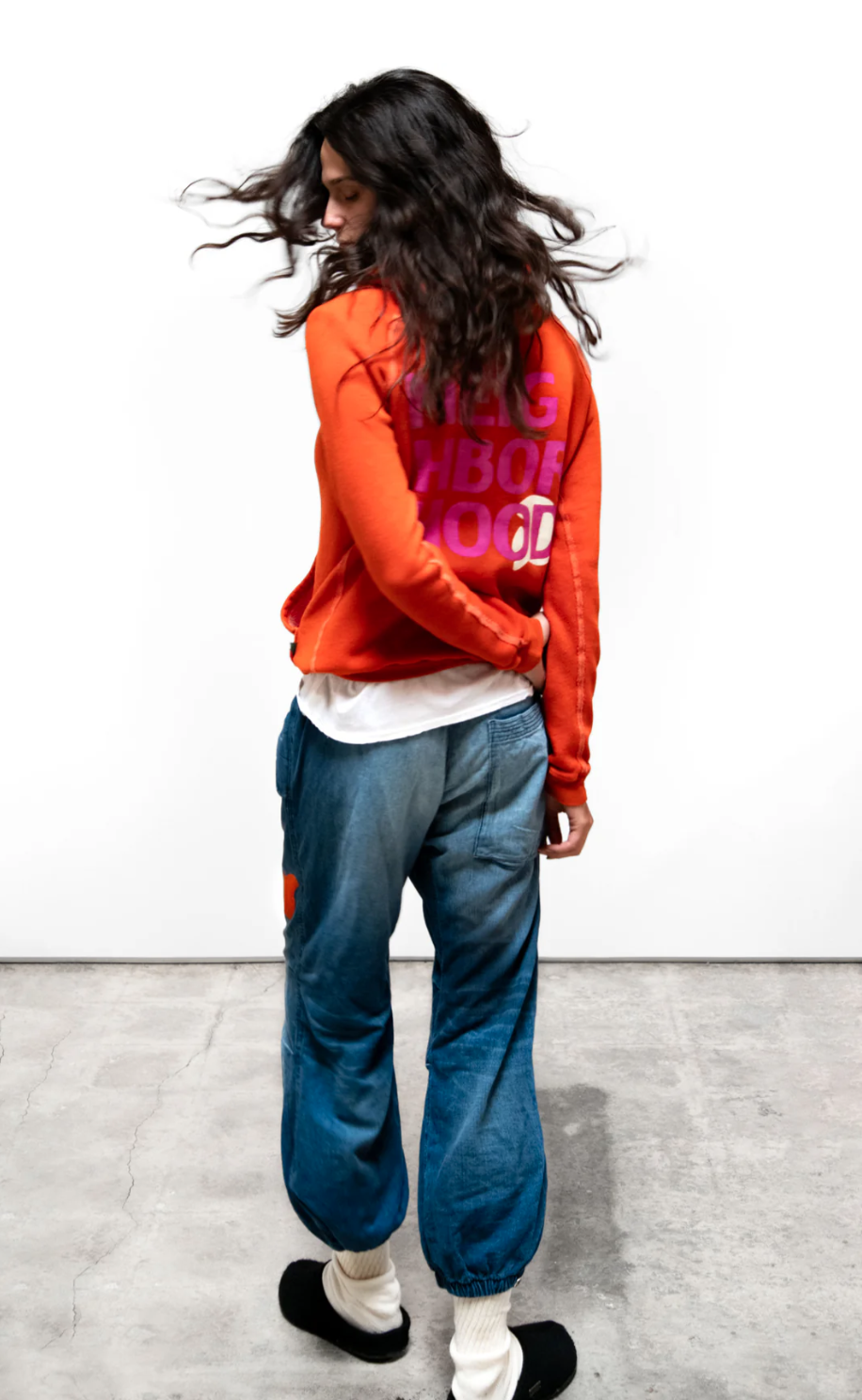 A person with long, wavy hair, wearing a bright orange Free City (sparrow, LLC) cir99 NEIGHBORHOOD LETS GO Hoodie featuring pink text that reads "FREECITYDOVE," loose blue jeans, white socks, and black slippers stands on a concrete floor against a white background. The person is facing away and appears to be in mid-motion, with hair flowing.