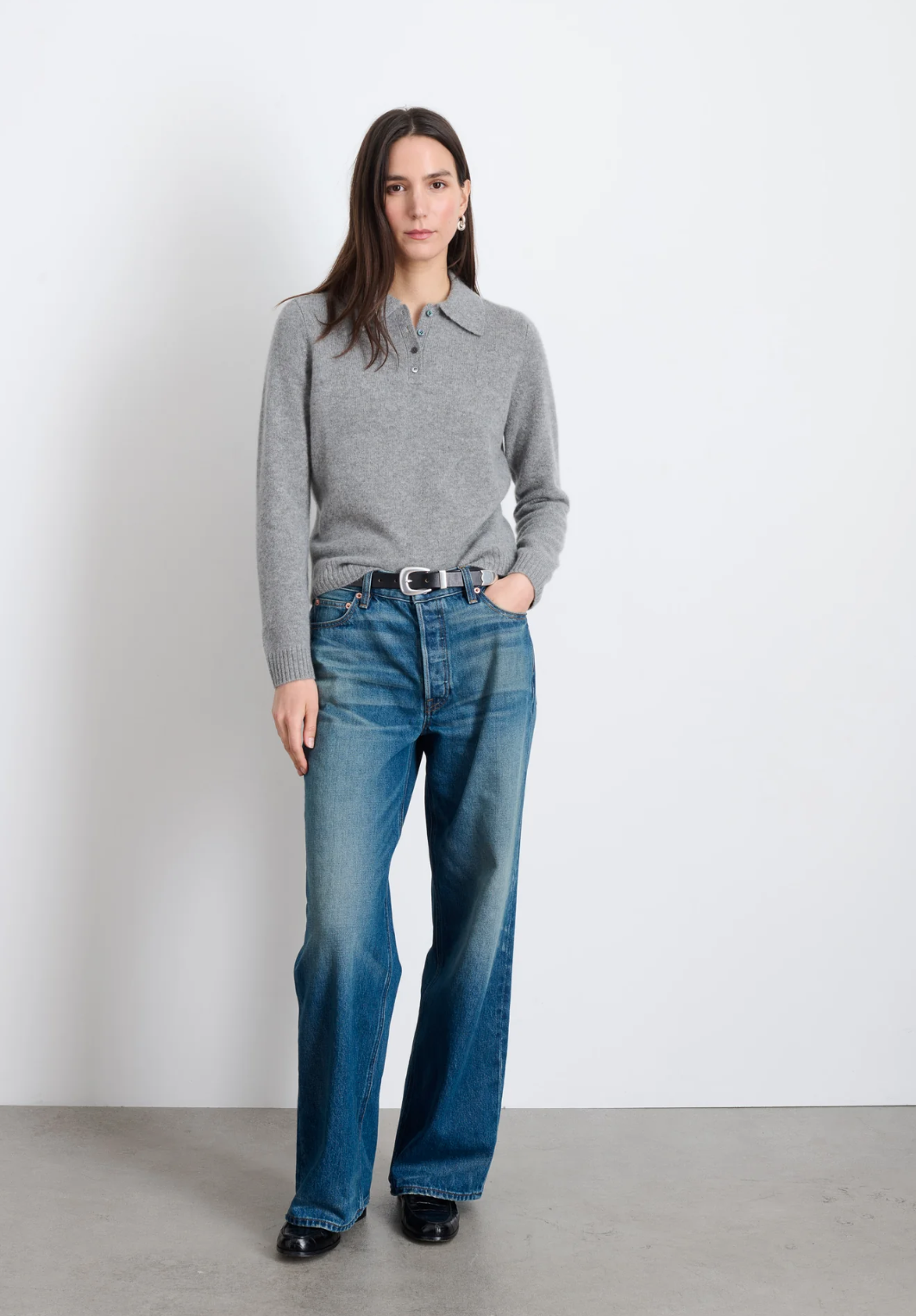 A woman with long hair stands against a plain white backdrop. She is wearing an Alex Mill Cashmere Alice Polo Sweater in gray, blue high-waisted wide-leg jeans, and black shoes. Her left hand is in her pocket, and she has a neutral expression. The extra softness of the sweater adds to her relaxed demeanor.