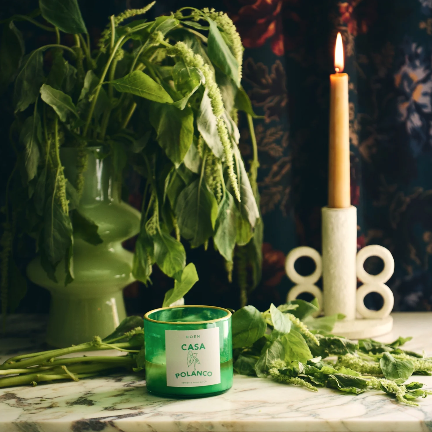 A green vase filled with lush, leafy plants is placed on a marble table. Beside it, a lit beige candle in a unique circular holder shares the space with a green glass "Casa Polanco" candle from Faire's ROEN Mexico City Candles collection. The background features a dark, patterned fabric.