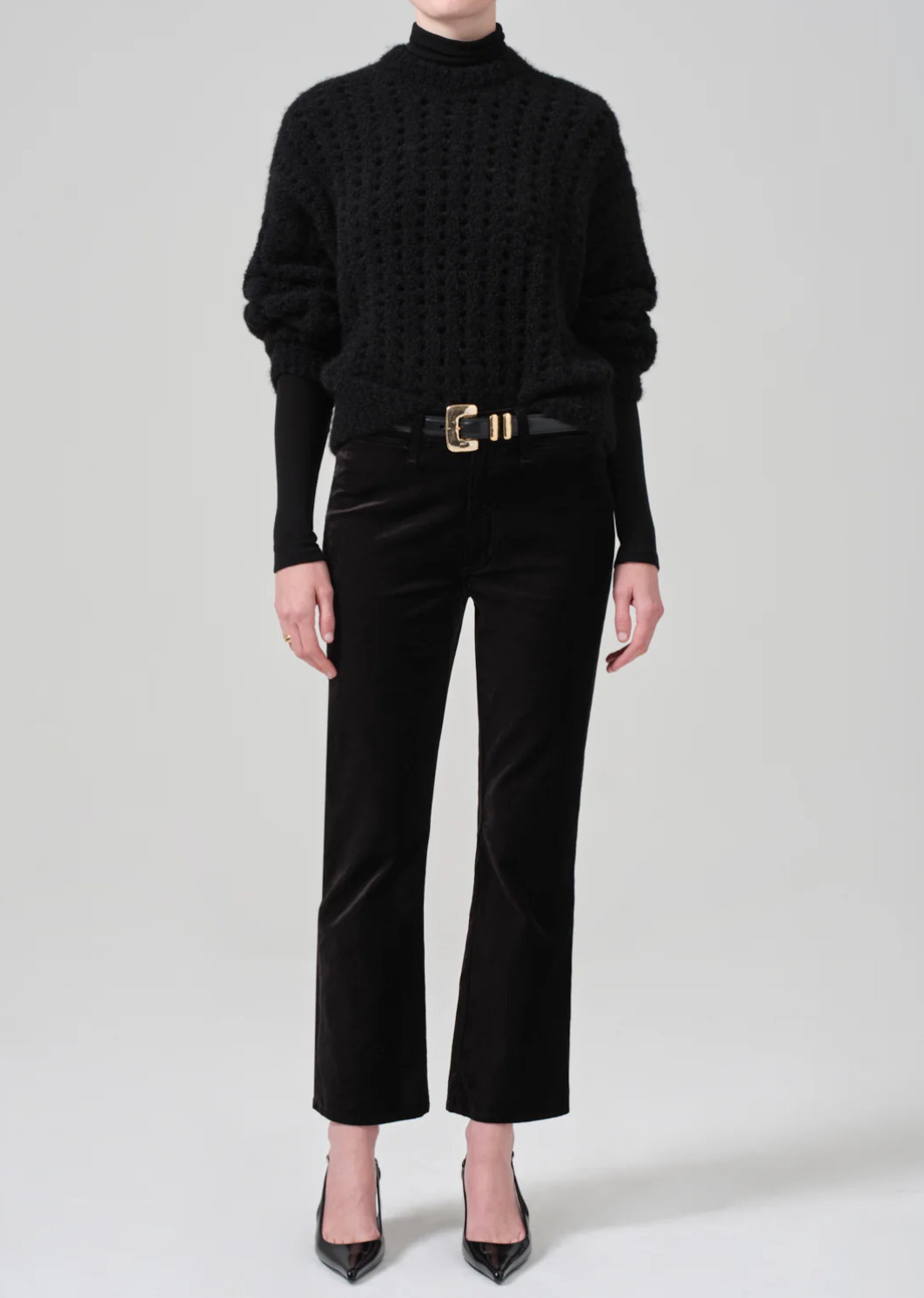 A person is elegantly dressed in a black textured high neckline sweater, paired with the Isola Cropped Trouser in Velvet from Citizens Of Humanity/AGOLDE, accented with a gold buckle belt, and sporting pointed black high heels. The backdrop is a plain white, creating a chic and sophisticated look.