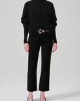 A person is elegantly dressed in a black textured high neckline sweater, paired with the Isola Cropped Trouser in Velvet from Citizens Of Humanity/AGOLDE, accented with a gold buckle belt, and sporting pointed black high heels. The backdrop is a plain white, creating a chic and sophisticated look.