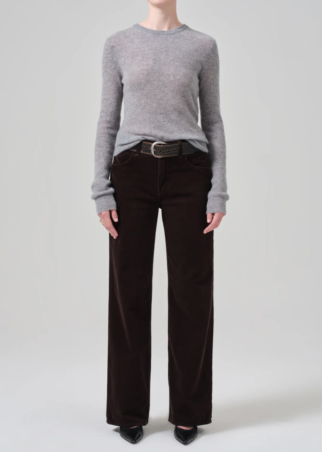 A person is wearing a light gray long-sleeve knit sweater, tucked into the Annina 30 Zip Fly In Corduroy pants by Citizens Of Humanity/AGOLDE. The high-waisted, dark brown wide-leg corduroy pants are held up with a textured belt. They are standing against a plain light gray background, wearing black pointed-toe shoes. The person&#39;s face is not visible.