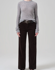 A person is wearing a light gray long-sleeve knit sweater, tucked into the Annina 30 Zip Fly In Corduroy pants by Citizens Of Humanity/AGOLDE. The high-waisted, dark brown wide-leg corduroy pants are held up with a textured belt. They are standing against a plain light gray background, wearing black pointed-toe shoes. The person's face is not visible.