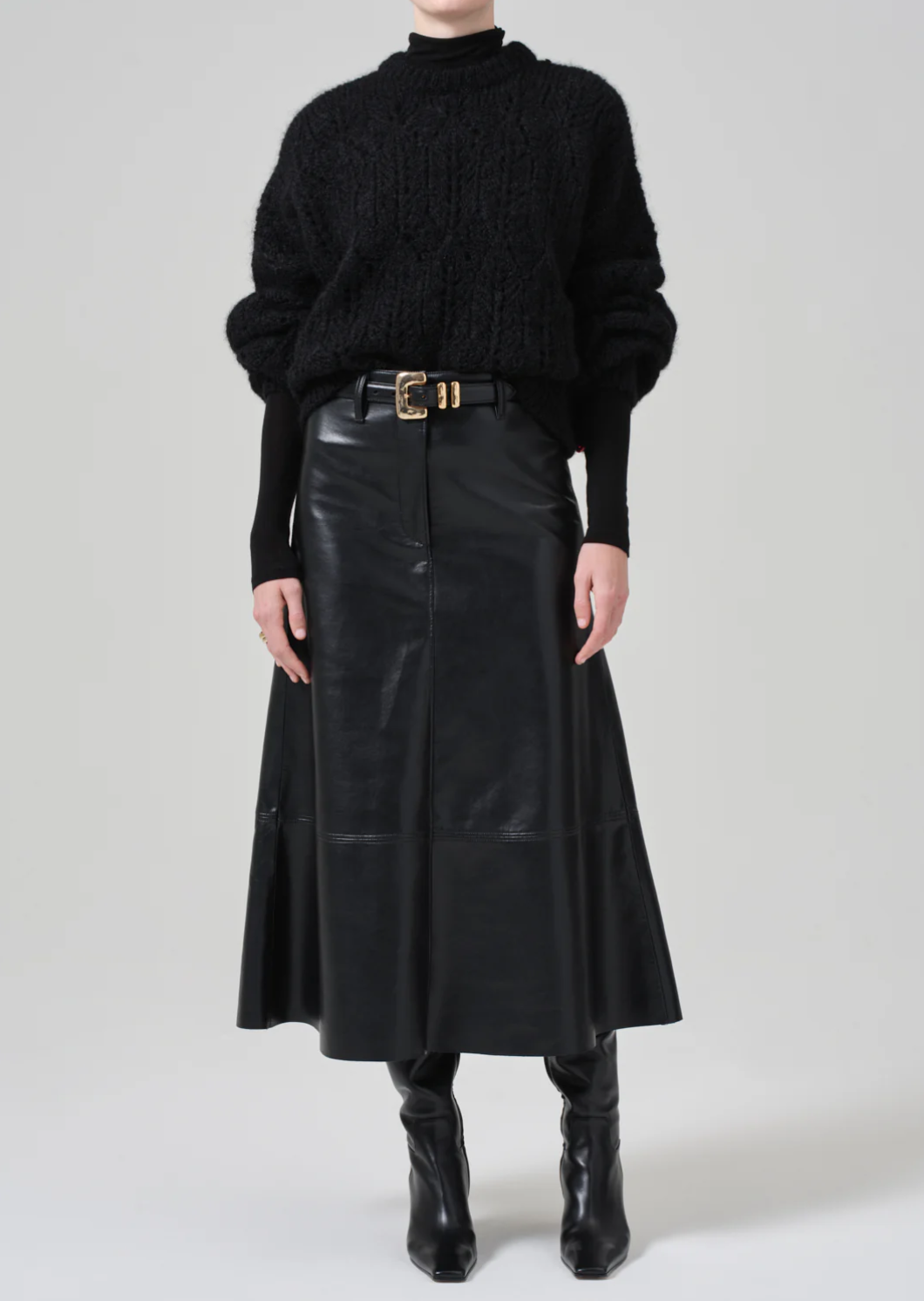 A person is wearing a chunky black turtleneck sweater, the Cassia Leather Skirt by Citizens Of Humanity/AGOLDE—a long high-rise recycled leather skirt with a belt featuring a gold buckle—and black leather boots. The outfit is shown against a plain gray background.