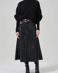 A person is wearing a chunky black turtleneck sweater, the Cassia Leather Skirt by Citizens Of Humanity/AGOLDE—a long high-rise recycled leather skirt with a belt featuring a gold buckle—and black leather boots. The outfit is shown against a plain gray background.