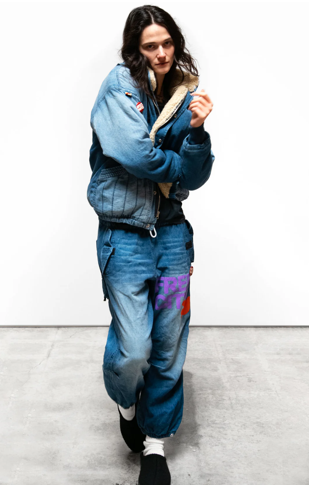 A person with long hair is posing casually against a white background. They are wearing a blue denim jacket with fur lining and the FLAP/SNAP SHELL FREECITY INDIGO pant - INDIGO2 from Free City (sparrow, LLC), which features colorful patches including the FREECITY print. With one hand raised, their expression remains neutral.