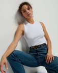 A person with curly hair is wearing the Joan Sleeveless Mock Neck by Nation LTD and high-waisted blue jeans with a black belt. They are sitting against a wall, legs apart, and looking thoughtfully at the camera.