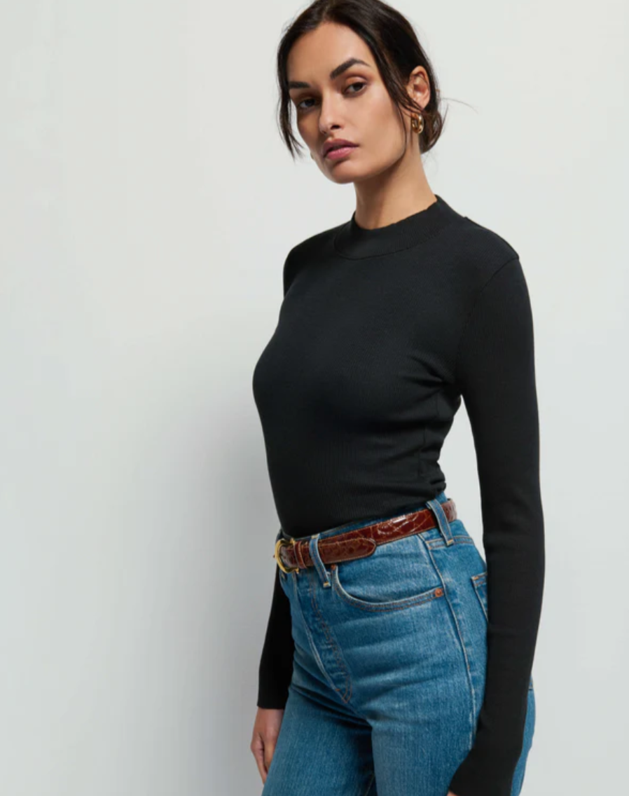 A person with dark hair tied back is wearing the Gina Slim Mock Neck by Nation LTD in jet black, paired with blue jeans and a brown belt. They are standing against a plain white background, looking directly at the camera with a neutral expression.