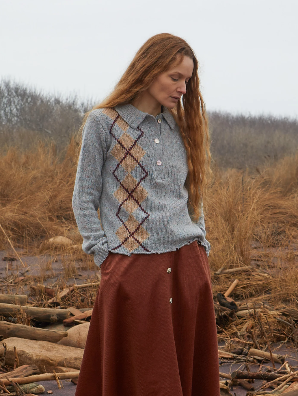 A person with long, wavy red hair stands outdoors in an overcast setting. They are wearing a Lee Tweed Argyle Polo Shirt by Lingua Franca and a red-brown skirt. The background features dried grass and barren trees, creating a natural, rustic atmosphere.