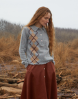 A person with long, wavy red hair stands outdoors in an overcast setting. They are wearing a Lee Tweed Argyle Polo Shirt by Lingua Franca and a red-brown skirt. The background features dried grass and barren trees, creating a natural, rustic atmosphere.