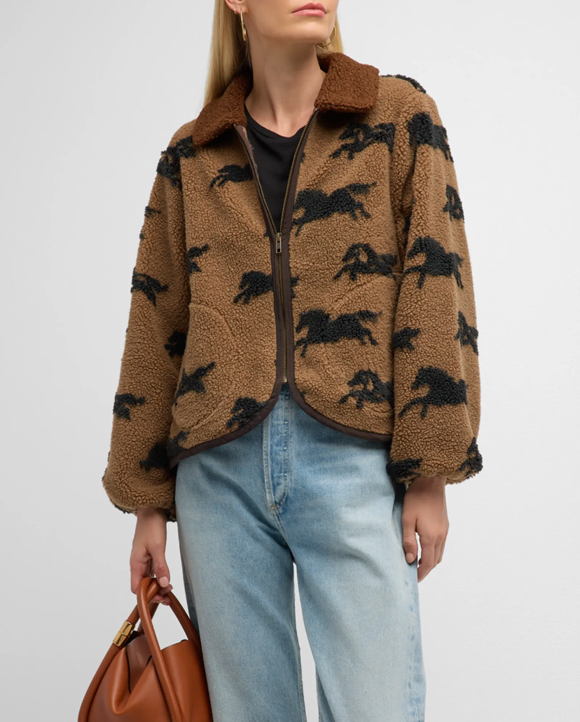 A person wearing The Great Inc.'s "The Pasture Jacket," a brown and black patterned, relaxed-fit teddy jacket with a horse motif print, light blue jeans, and a black top holds a brown leather handbag. The jacket features a zip-up front and a large collar. The person has blonde hair, and the background is plain white.