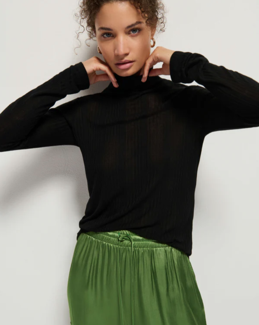 A person with curly hair is posing in the Rene Turtleneck by Nation LTD. This long-sleeved black turtleneck features ribbed details and is paired with green satin pants. The individual stands against a plain white background, their hands near their neck as they look directly at the camera.