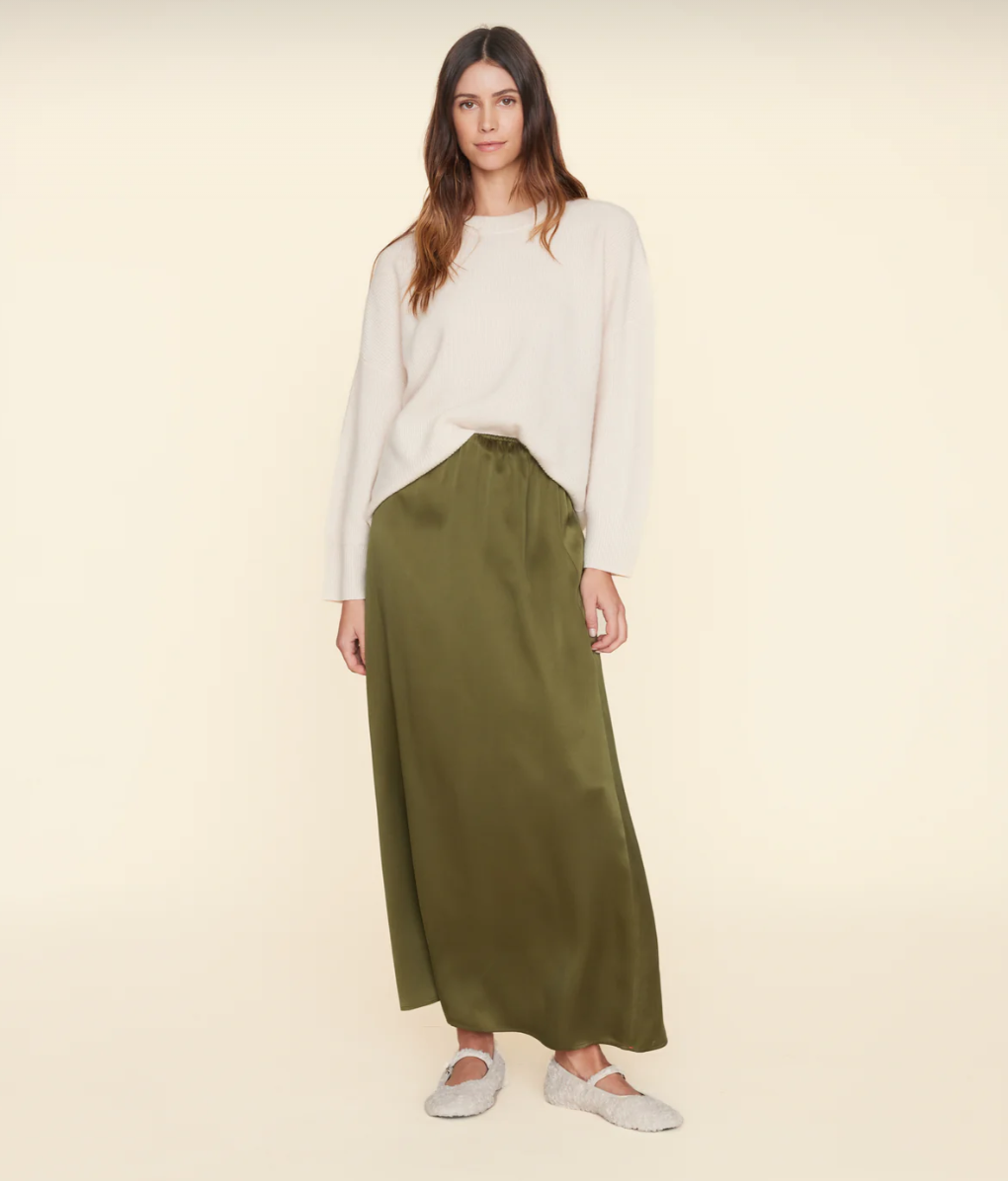 The Sela Skirt by Xirena, a midi-length, olive-green piece with a straight cut, is shown against a plain beige background. Made of luxurious silk charmeuse and featuring an elastic waistband, this slip-on skirt exudes effortless elegance.