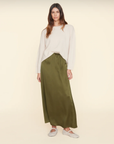 The Sela Skirt by Xirena, a midi-length, olive-green piece with a straight cut, is shown against a plain beige background. Made of luxurious silk charmeuse and featuring an elastic waistband, this slip-on skirt exudes effortless elegance.