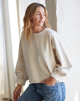 A woman with wavy, light brown hair is wearing the **Zayne Terry Pullover** by **Perfectwhitetee**, a loose beige sweatshirt with pintucked wrists, and blue jeans. She is leaning against a textured white wall and looking to her right. Light filters through sheer curtains in the background, highlighting her relaxed silhouette.