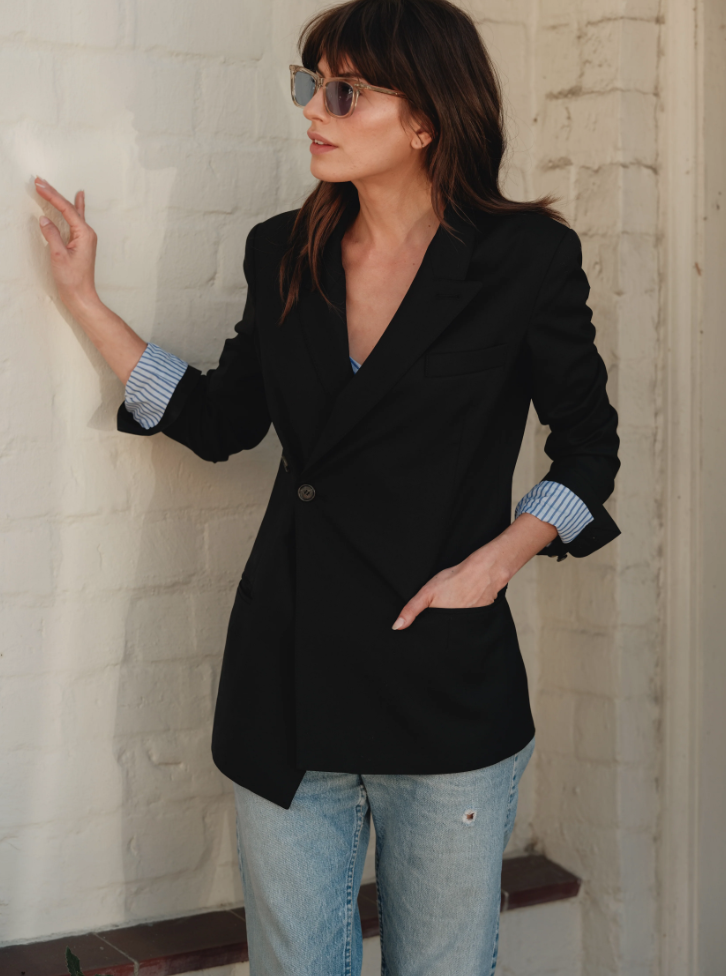 A woman wearing stylish sunglasses and the Cissa X SMYTHE Blazer over a light blue wrap shirt stands against a white brick wall. She has one hand in her jean pocket and the other hand lightly touching the wall, looking to the side.