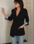 A woman wearing stylish sunglasses and the Cissa X SMYTHE Blazer over a light blue wrap shirt stands against a white brick wall. She has one hand in her jean pocket and the other hand lightly touching the wall, looking to the side.