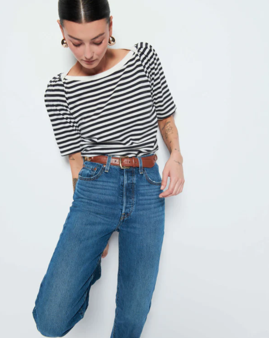 A person stands against a plain background, looking down. They are wearing a Deana Stripe Envelope T-Shirt in black and white from Nation LTD, high-waisted blue jeans, and a brown belt. The individual has short, dark hair and gold hoop earrings, with visible tattoos on their forearms.