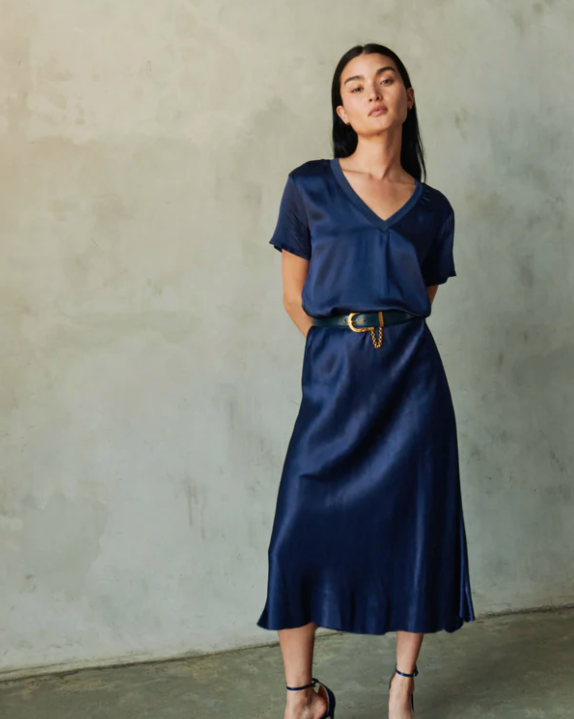 A person with long dark hair is standing against a concrete wall, wearing a navy blue silk blouse tucked into a Nation LTD's Mabel Bias Skirt. They have a black and gold belt and black sandals, with their hands behind their back.