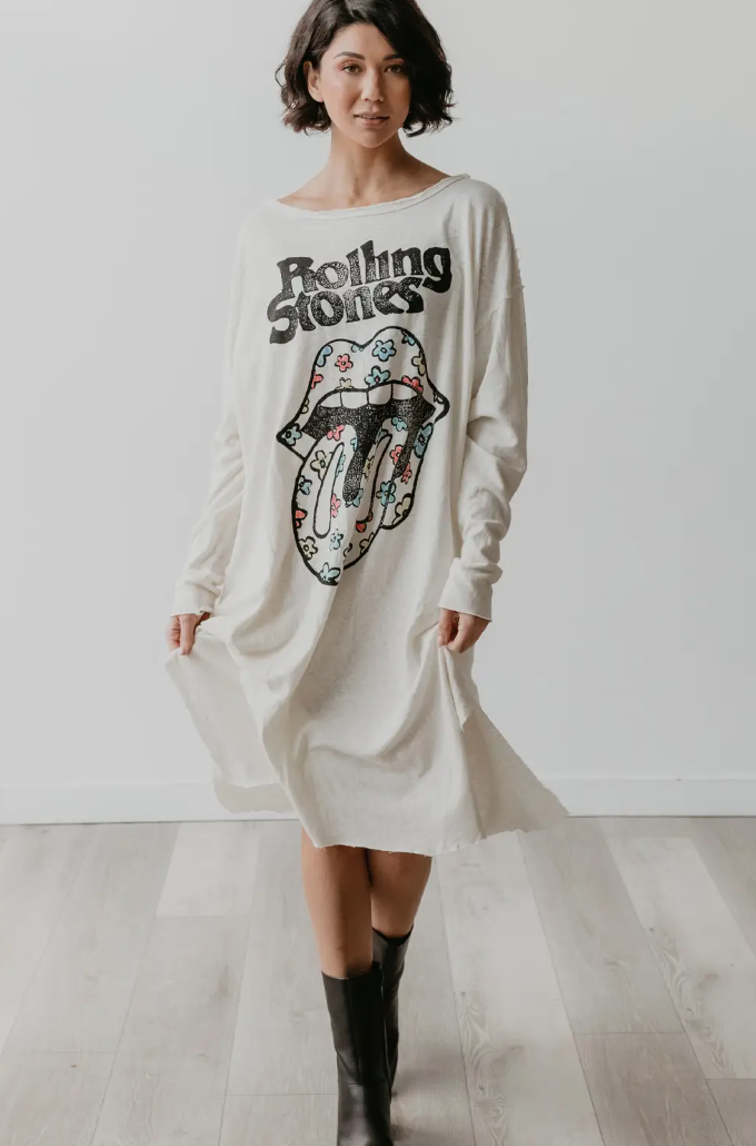 A woman stands indoors wearing the Flower Power Stones Tee Dress by Faire, which features an ethically made Rolling Stones tongue and lips logo adorned with colorful patterns. She pairs the long white dress with black knee-high boots, creating a chic contrast against the simple, plain white wall and wooden floor backdrop.