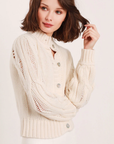 A woman with shoulder-length brown hair is wearing a cream-colored Ava Ladder Cable Crop Cardi from Feller, featuring Natural Shell Buttons down the front. She is looking slightly to the right and holding a strand of her hair with her left hand. The background is plain white. This sweater, available in size S, should be hand washed.