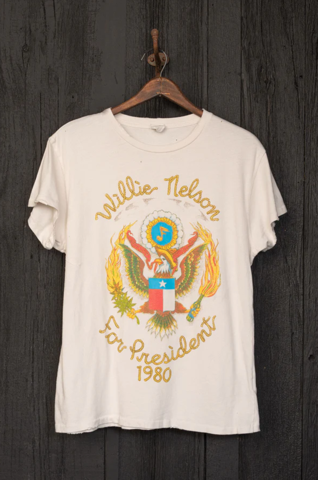 The Willie Nelson For President Tee by Made Worn, a white T-shirt made in the USA, is displayed on a wooden hanger. It showcases a vibrant graphic of an eagle with outstretched wings, accompanied by the text "Willie Nelson" above and "For President 1980" below. This vintage unisex fit shirt stands out against a dark wooden wall background.