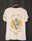 The Willie Nelson For President Tee by Made Worn, a white T-shirt made in the USA, is displayed on a wooden hanger. It showcases a vibrant graphic of an eagle with outstretched wings, accompanied by the text "Willie Nelson" above and "For President 1980" below. This vintage unisex fit shirt stands out against a dark wooden wall background.