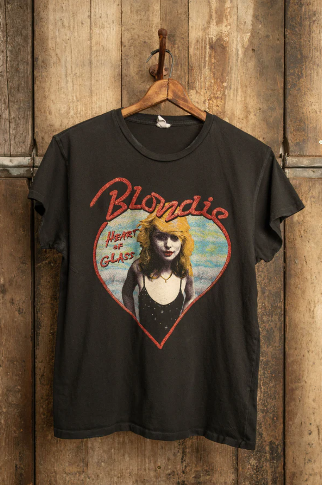 The Blondie Tee by Made Worn is a black, vintage-style unisex shirt featuring a graphic of a woman inside a red heart with "Blondie" written above. Below the heart, the text reads "Heart of Glass." Made in the USA, this tee is displayed on a wooden hanger against a rustic wooden background.