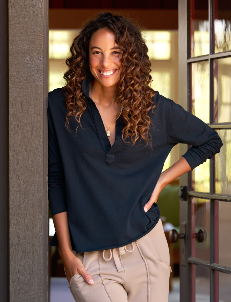 A smiling woman with curly hair and hands in her pockets leans against a door frame. She is wearing the Patrick Henley Jersey by Frank & Eileen, a dark preppy tee made of 100% cotton, paired with light-colored pants in a cropped relaxed fit. The background includes a door with glass panes and a softly lit interior space.