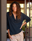 A smiling woman with curly hair and hands in her pockets leans against a door frame. She is wearing the Patrick Henley Jersey by Frank & Eileen, a dark preppy tee made of 100% cotton, paired with light-colored pants in a cropped relaxed fit. The background includes a door with glass panes and a softly lit interior space.