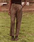 A person standing on grass wears The Maker Trouser, a pair of high-waisted, pleated wool blend trousers from The Great Inc., paired with a black top. With one hand in their pocket, black ankle boots complete the look. The background features a fenced area, possibly a farm, and a grassy field, evoking timeless vintage menswear vibes.