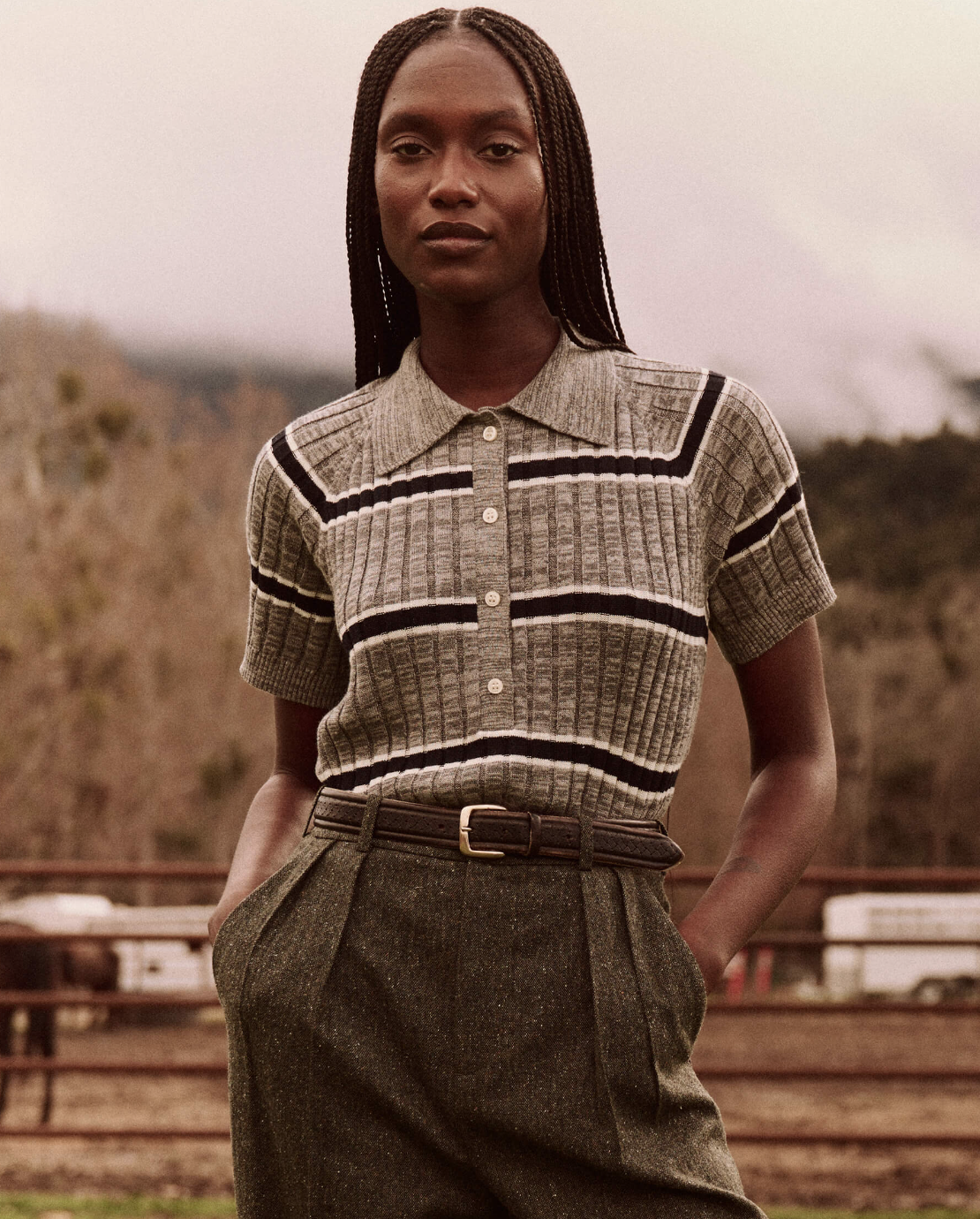 A person with long braided hair is wearing The Great Inc.'s Classic Polo Sweater in heather grey, which features short sleeves, a fine-gauge ribbed knit, and striped patterns. They have paired it with high-waisted trousers and a belt. They are standing outdoors with a blurred background showcasing trees and a wooden fence.