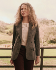 A person with curly hair stands outdoors in front of a wooden fence, surrounded by a tree-filled landscape. They are wearing The Double Breasted Blazer from The Great Inc. over a white, textured blouse and burgundy pants. The atmosphere is serene and rural.