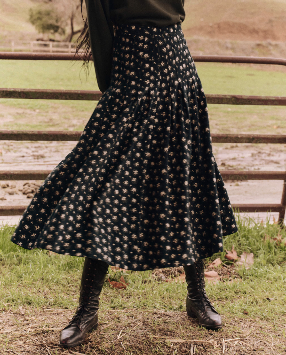 A person wearing The Great Inc.'s The Rhythm Skirt featuring a Night Lily Print, complemented by black lace-up boots, stands in front of a wooden fence in a grassy area. The person's upper body is not visible.