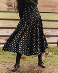 A person wearing The Great Inc.'s The Rhythm Skirt featuring a Night Lily Print, complemented by black lace-up boots, stands in front of a wooden fence in a grassy area. The person's upper body is not visible.