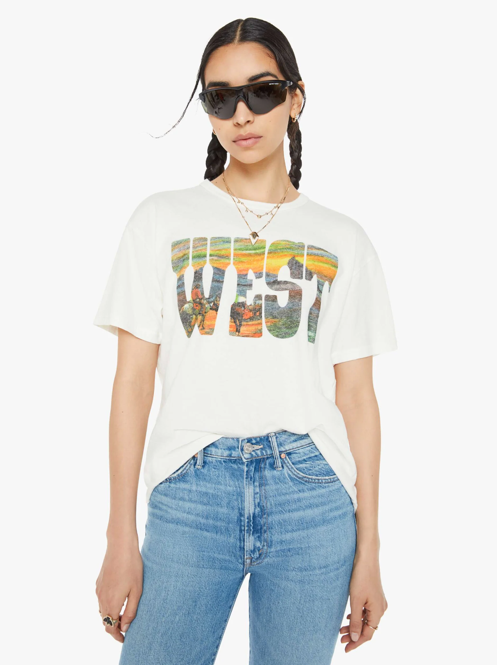 A person with braided hair is sporting The Sleep Over, an oversized white T-shirt by Mother, crafted from 100% cotton. This shirt prominently features the word "WEST" in large letters, along with a Western sunset image. They're also wearing blue jeans and layered necklaces, standing against a plain background while accessorized with sunglasses.