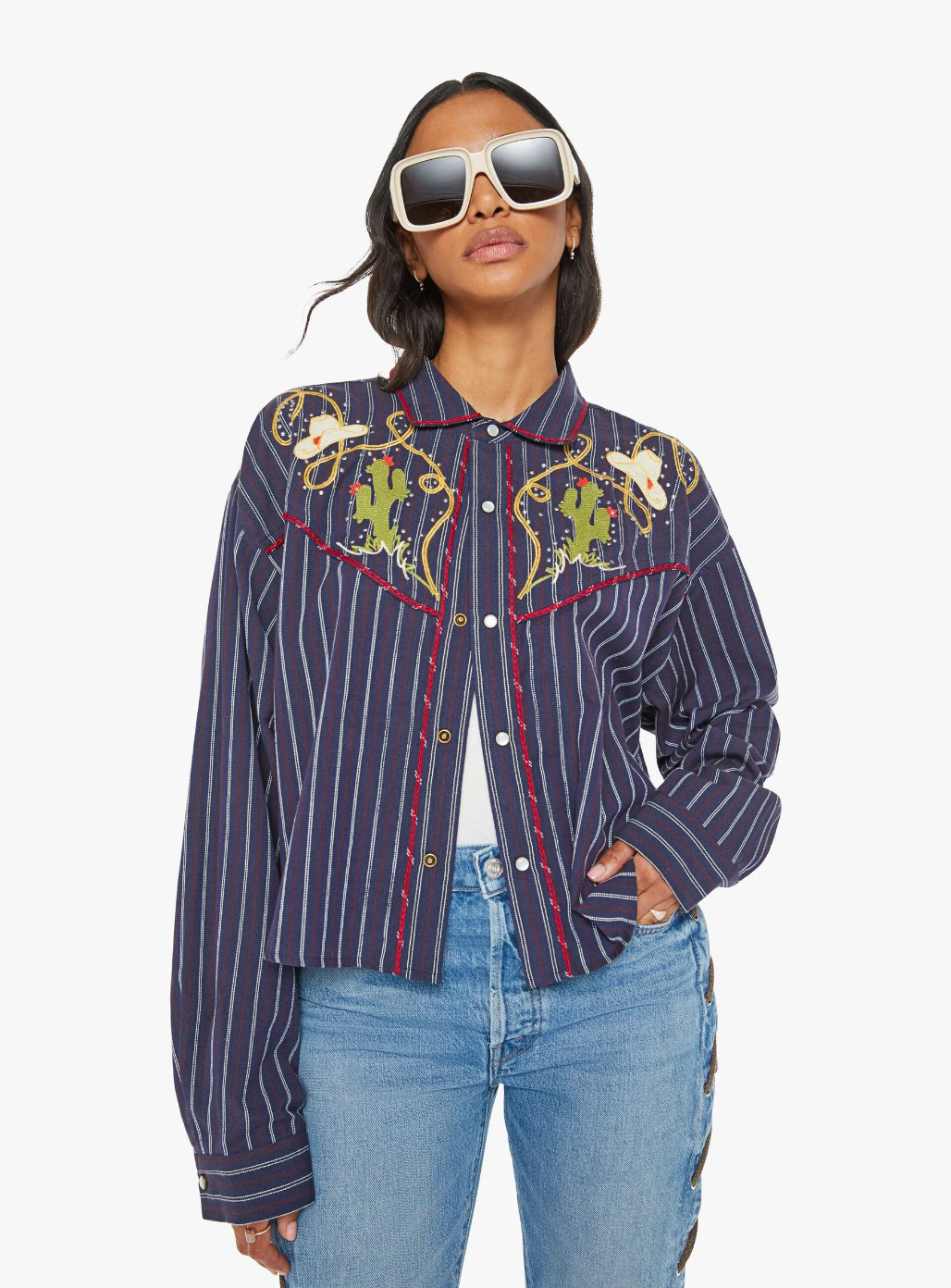 A person sporting the stylish "The Riding Shotgun" jacket by Mother, featuring blue stripes and cowboy embroidery, paired with light blue jeans. They have long hair and wear large, square sunglasses. Their hand rests confidently in a pocket as they pose against a plain background.