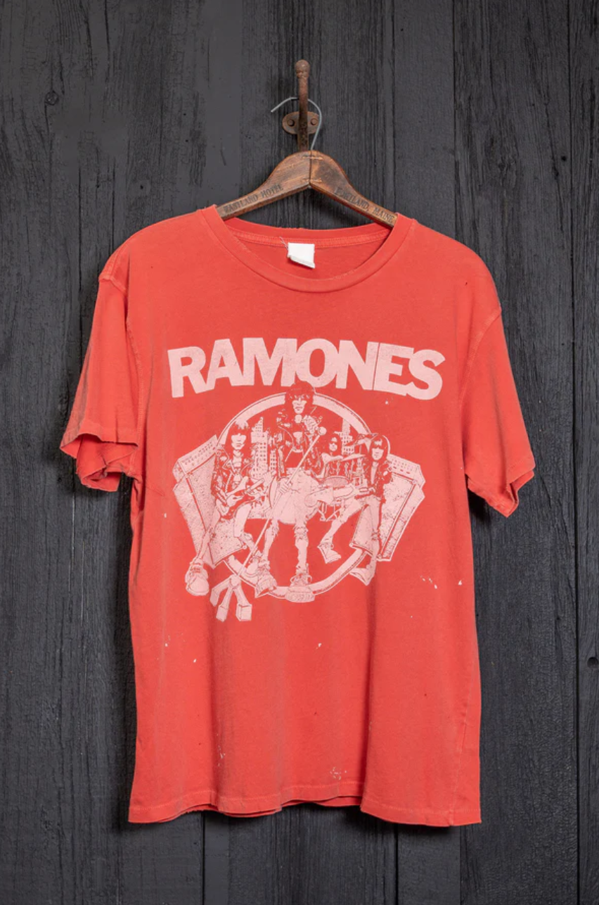 The Ramones Crew Tee by Made Worn is a red cotton T-shirt displaying unique distressing and a white graphic of four cartoon figures, along with "RAMONES" in bold letters. This vintage unisex fit shirt is showcased on a classic wooden hanger against a dark wooden backdrop.