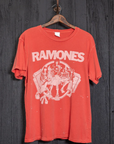 The Ramones Crew Tee by Made Worn is a red cotton T-shirt displaying unique distressing and a white graphic of four cartoon figures, along with "RAMONES" in bold letters. This vintage unisex fit shirt is showcased on a classic wooden hanger against a dark wooden backdrop.