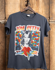 The Tom Petty 1979 Crew Tee by Made Worn is a vintage unisex fit black T-shirt showcasing a vibrant graphic design with hearts and a guitar. "Tom Petty and the Heartbreakers" encircles an illustrated portrait of a long-haired figure. Featuring unique distressing, it hangs against a wooden backdrop, exuding timeless charm.