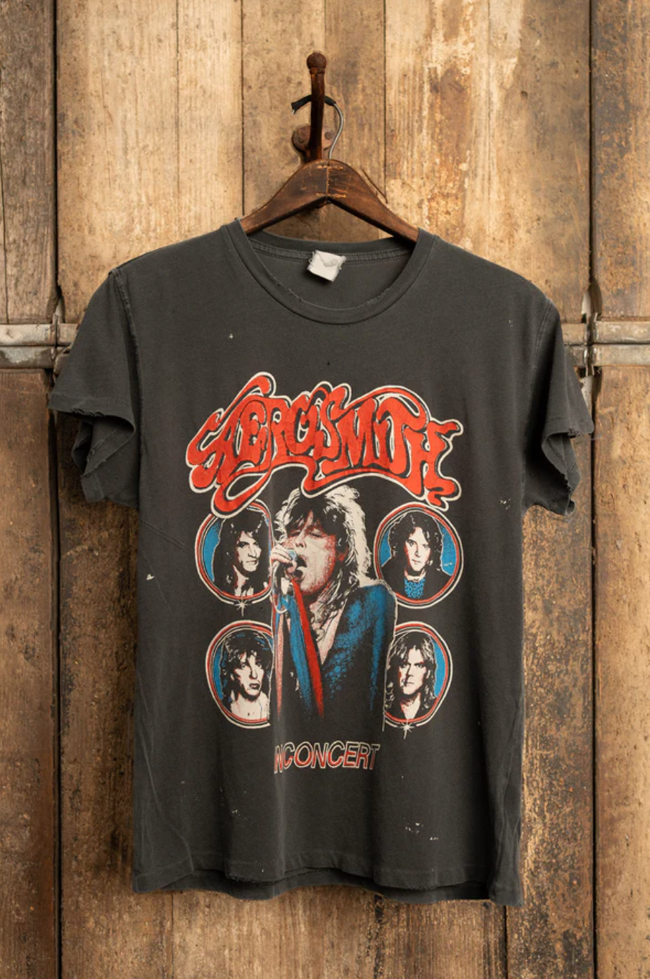 An "Aerosmith In Concert Crew Tee" from Made Worn hangs on a wooden door, featuring an eye-catching graphic bursting with vibrant illustrations of the band members and "IN CONCERT" in bold red. Handmade in Los Angeles, this unisex tee has a vintage cotton construction that exudes a charmingly worn, retro style.