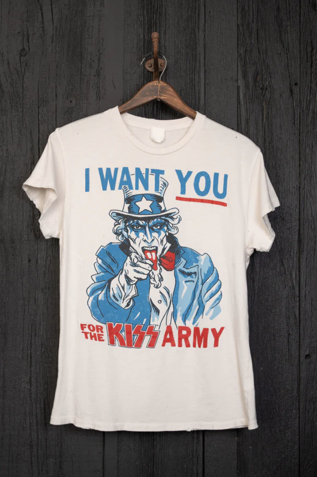 The Kiss Wants You Crew Tee by Made Worn, a vintage unisex white cotton t-shirt, is showcased on a wooden wall. Handmade in Los Angeles, it depicts Uncle Sam with face paint pointing forward and is adorned with the bold message "I WANT YOU FOR THE KISS ARMY" in striking blue and red letters.