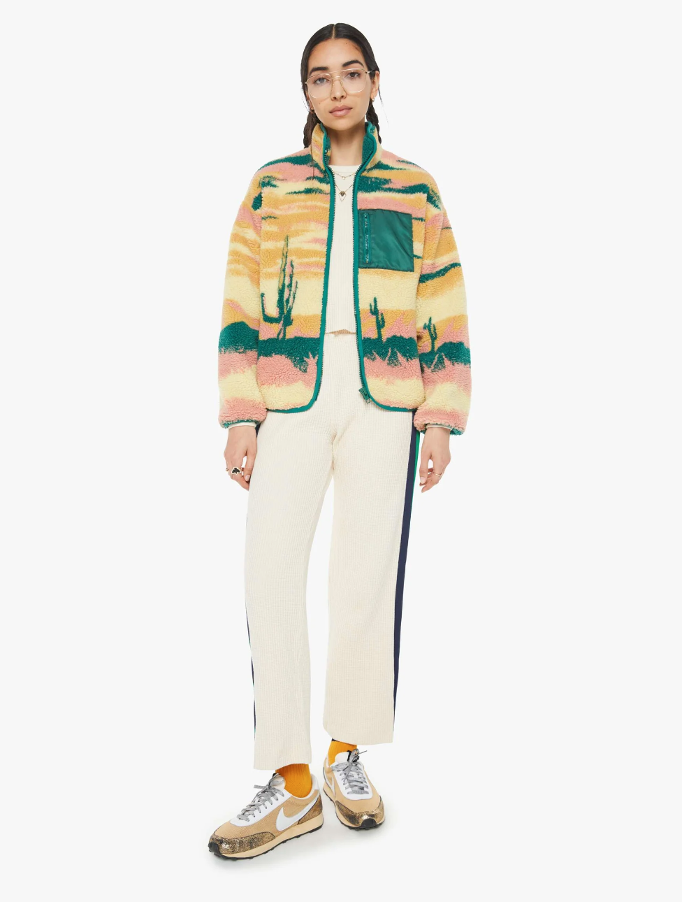 A person wearing glasses is styled in Mother's "The Heat Lamp Cactus Cooler" fleece jacket, featuring eye-catching desert sunset graphics and balloon sleeves. They combine it with white pants accented by side stripes, yellow socks, and gray sneakers. The jacket's cactus and sunset motifs pop against the clean white backdrop.